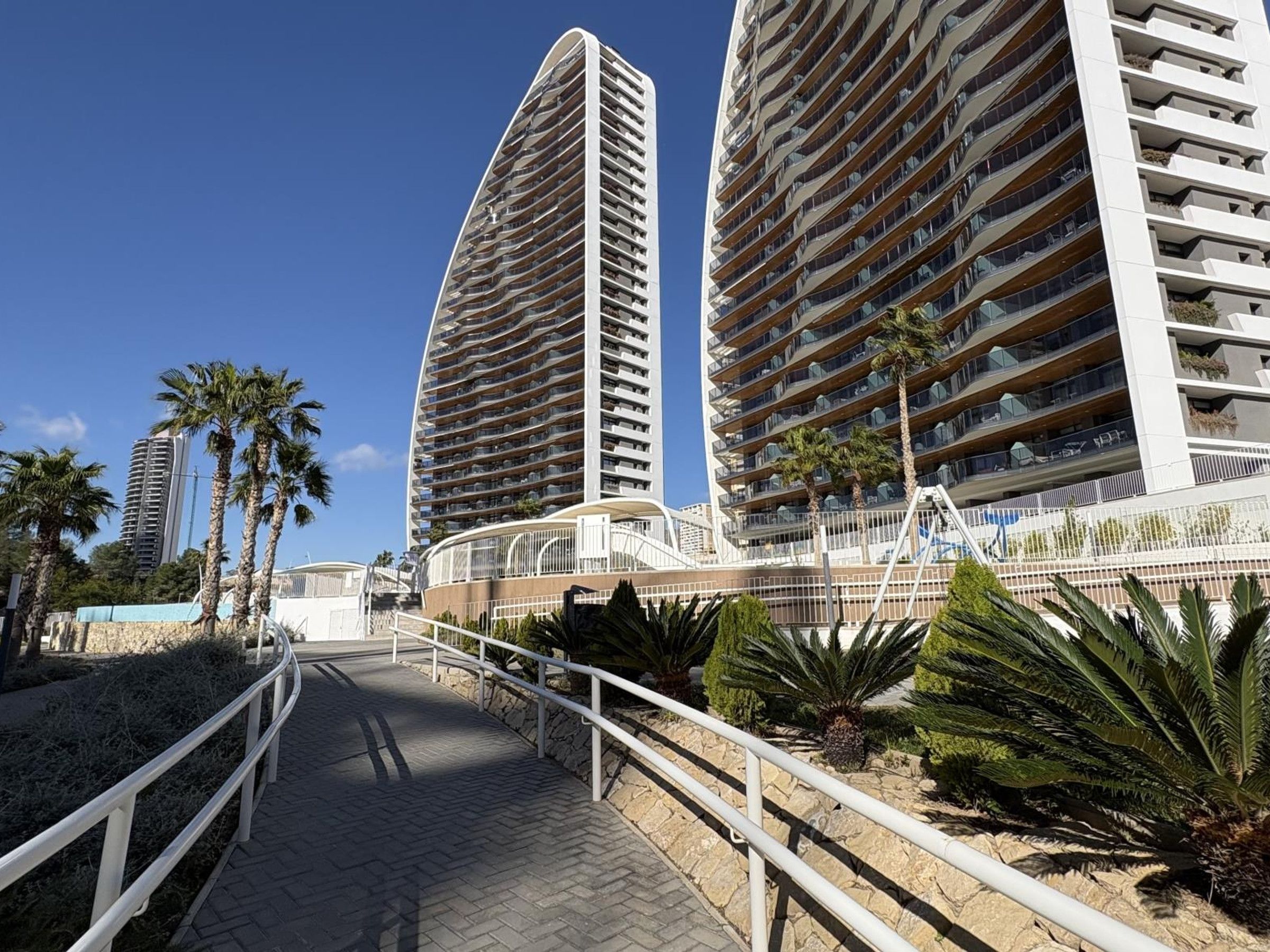 A pathway leading to a modern high-rise building with multiple balconies, surrounded by palm trees and a clear sky.