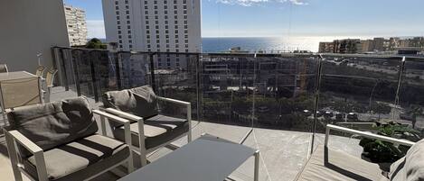 A balcony with a view of the sea and buildings, furnished with a sofa, chairs, and a table.