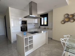 A modern kitchen with white cabinetry, a stainless steel range hood, a built-in oven, and a glass dining table with a white chair. - Spacious apartment, panoramic view - 6 guests (Benidorm)