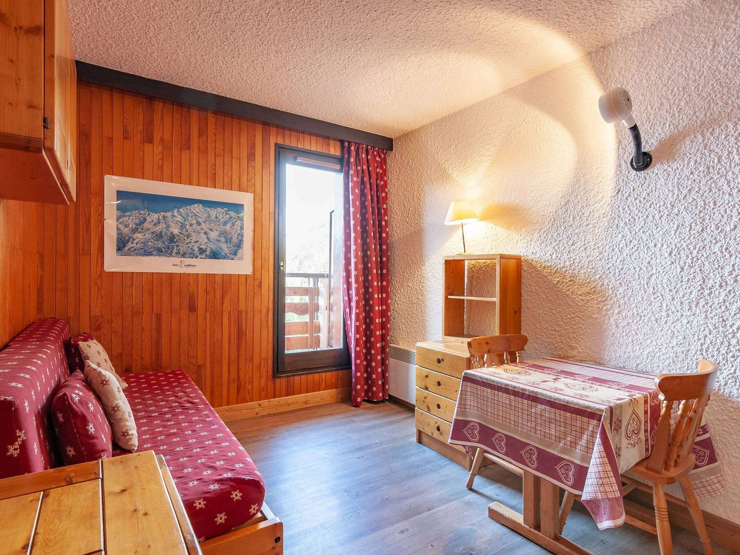 A cozy room with wooden walls, a bed with a red and white patterned bedspread, a wooden dresser, a table with a checkered tablecloth, and a window with curtains.