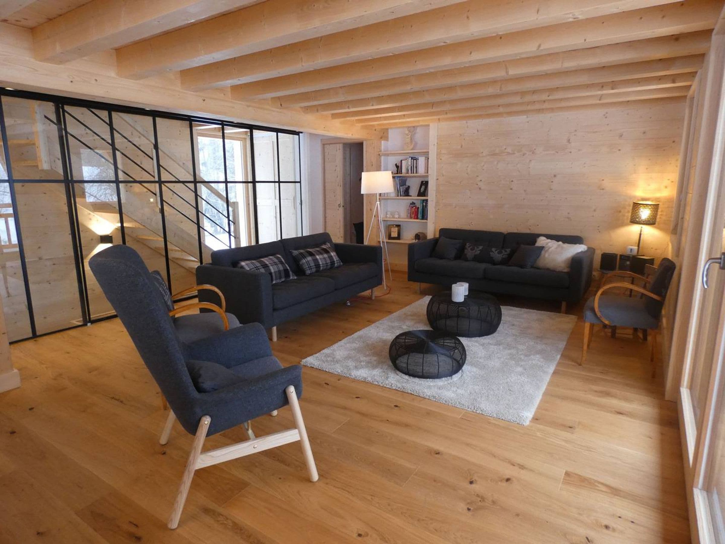 Ein modernes Wohnzimmer mit Sofa, Sessel und Couchtisch, Holzböden und einer Treppe mit Glastrennwand.