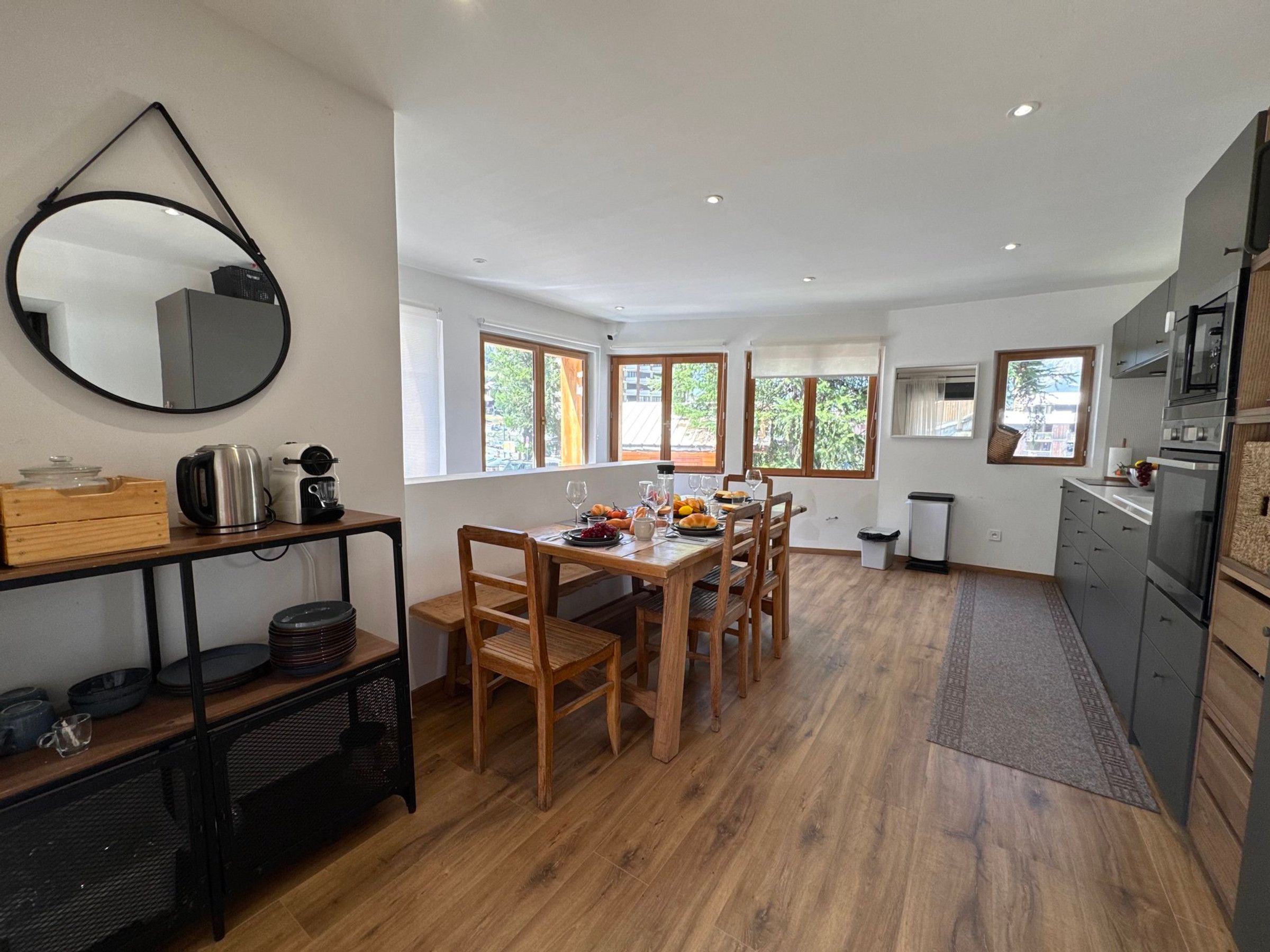 A modern kitchen with a dining area, wooden furniture, and large windows.