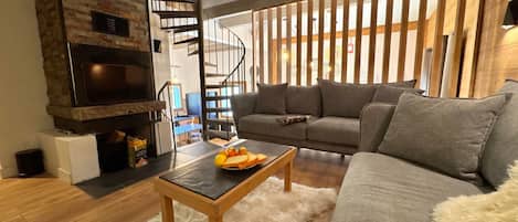 A cozy living room with a fireplace, a sofa, a coffee table with fruit, and a staircase.