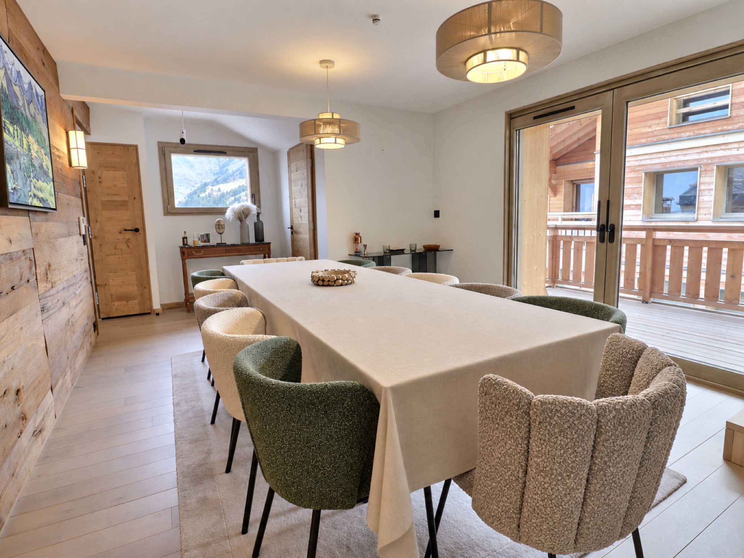 A dining area with a long table and chairs, a wooden wall, and a view through a glass door.