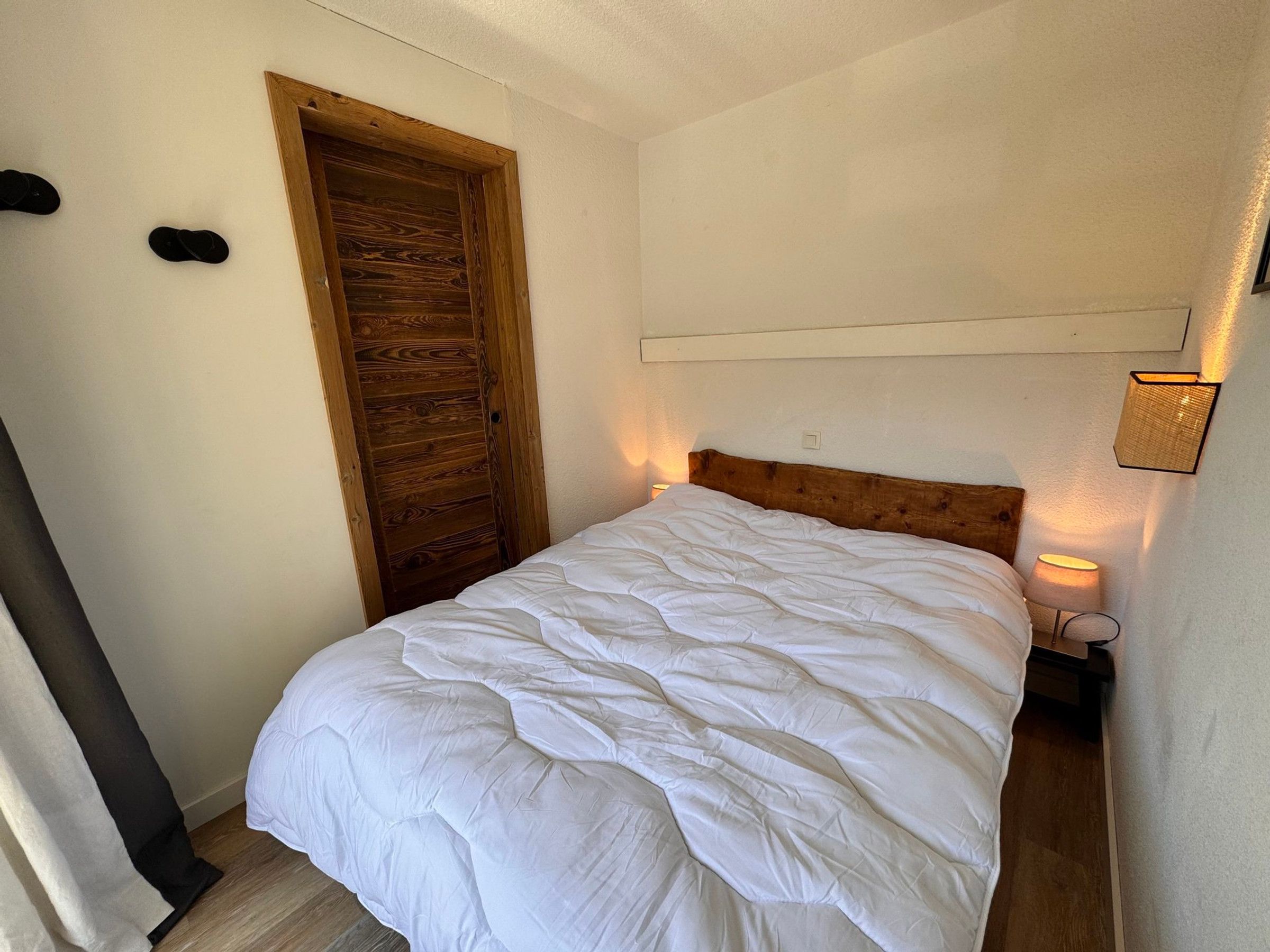 A bedroom with a wooden door, a bed with a white duvet, a bedside lamp, and a wall-mounted light fixture.