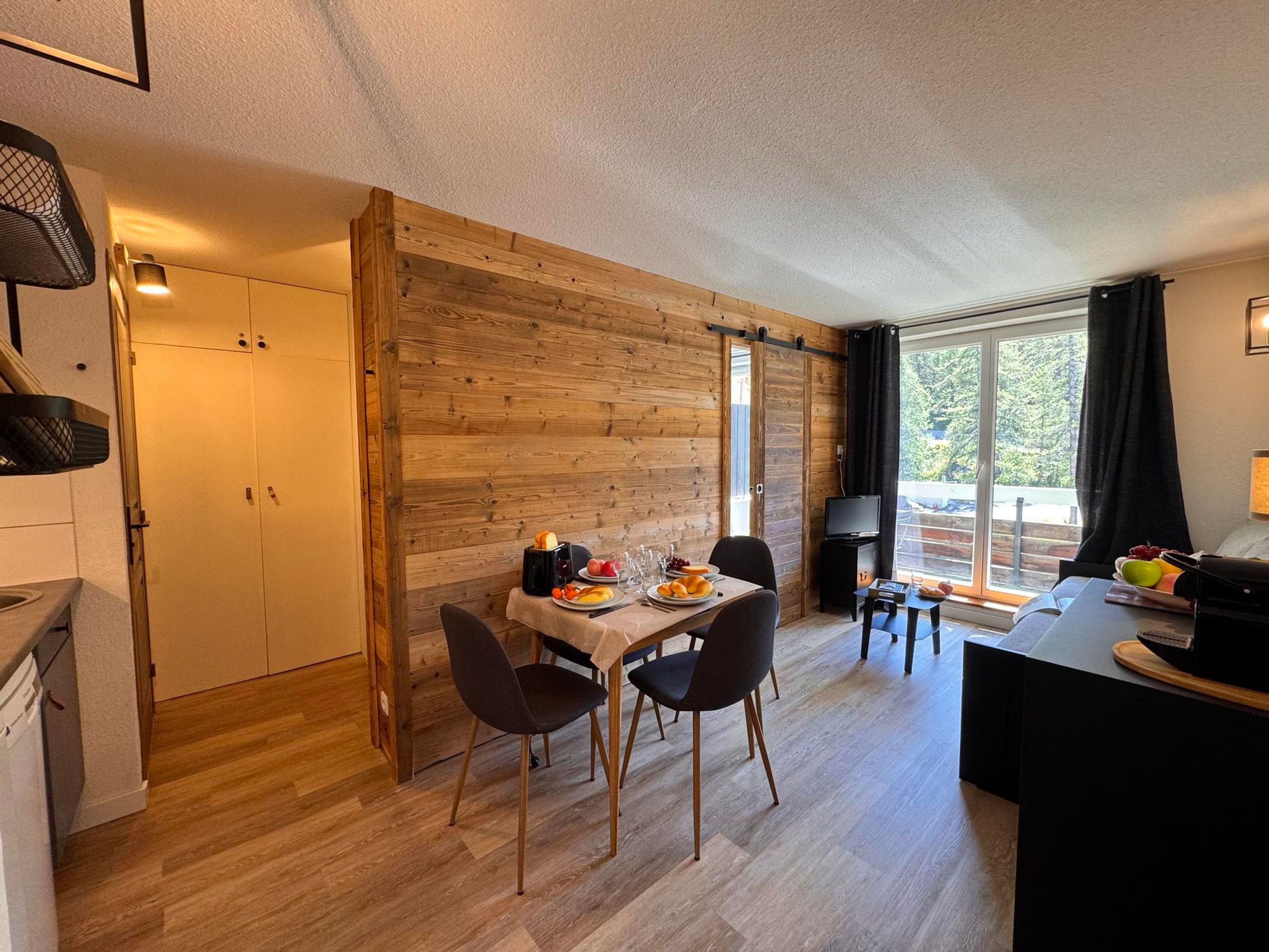 A compact room with a wooden wall, a dining table with chairs, a small TV, and a window with curtains.