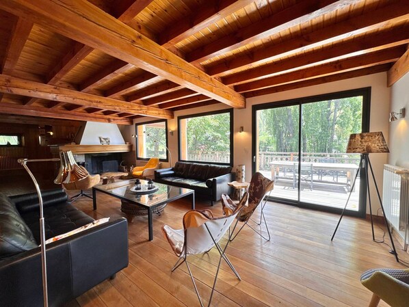 A spacious living area with a high ceiling, wooden beams, a fireplace, and large windows offering a view of the outdoors.