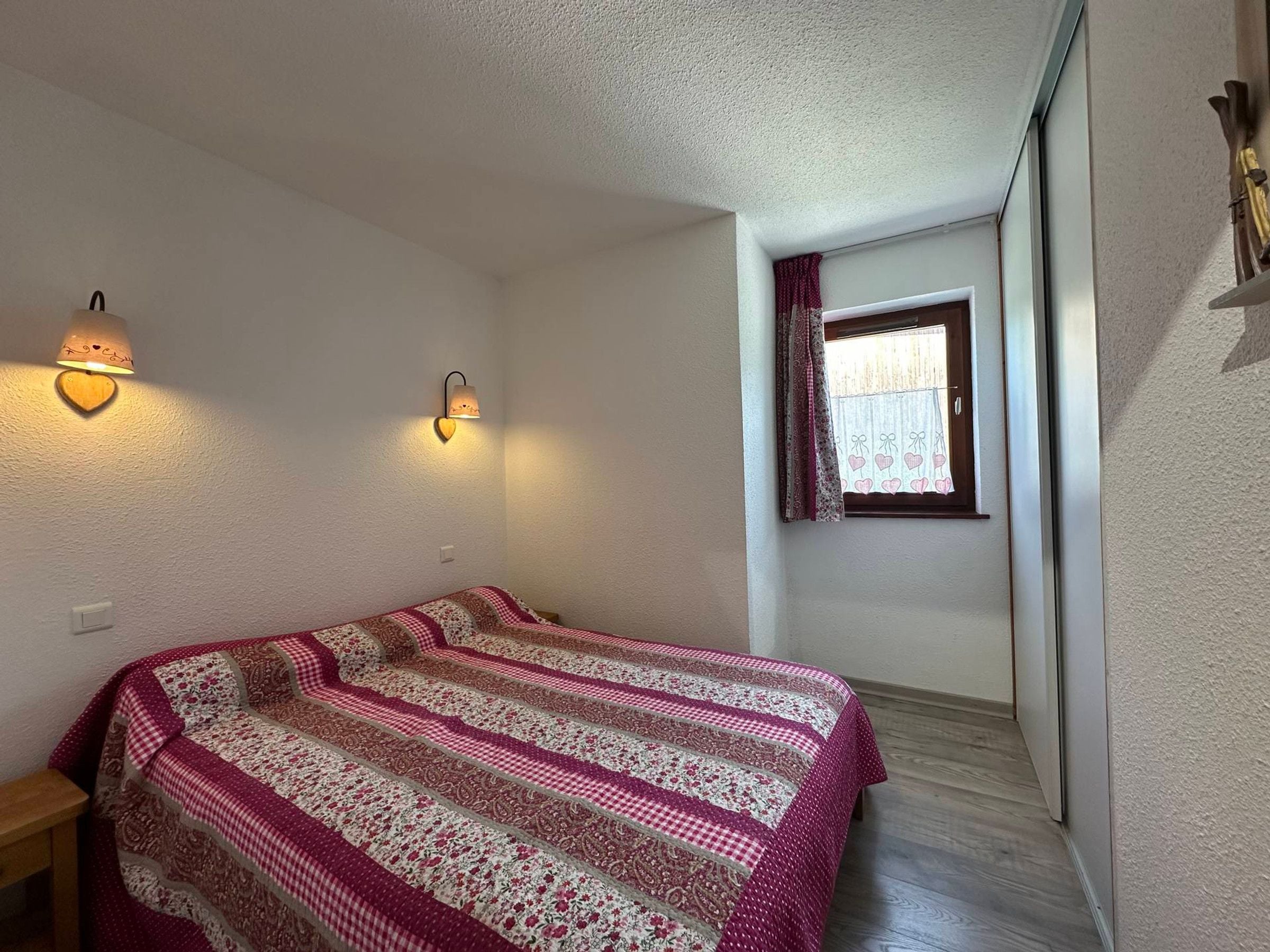 Un dormitorio con cama, dos lámparas de pared, una ventana con cortinas y suelo de madera.
