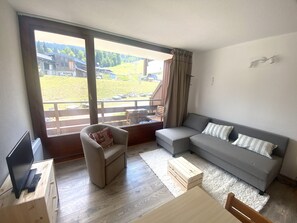 Ein modernes Wohnzimmer mit einem grauen Sofa, einem Holztisch und einem Sessel mit Kissen. Ein Laptop steht auf einem Holzständer. Große Fenster bieten Blick auf eine grüne Landschaft.