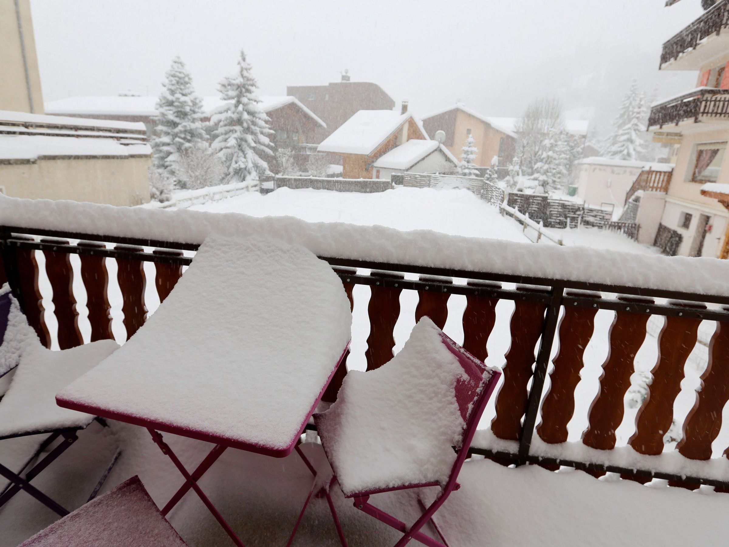 Un balcón con mesa y sillas cubiertas de nieve, con vista a una zona residencial con casas y árboles.