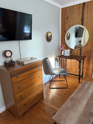 A room with a wooden dresser, a chair, a desk with a mirror, and a flat-screen TV mounted on the wall.