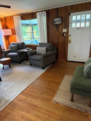 A cozy living room with two grey sofas, a small round table, and a green armchair. A wooden floor and a white door with a 'No Smoking' sign are visible.