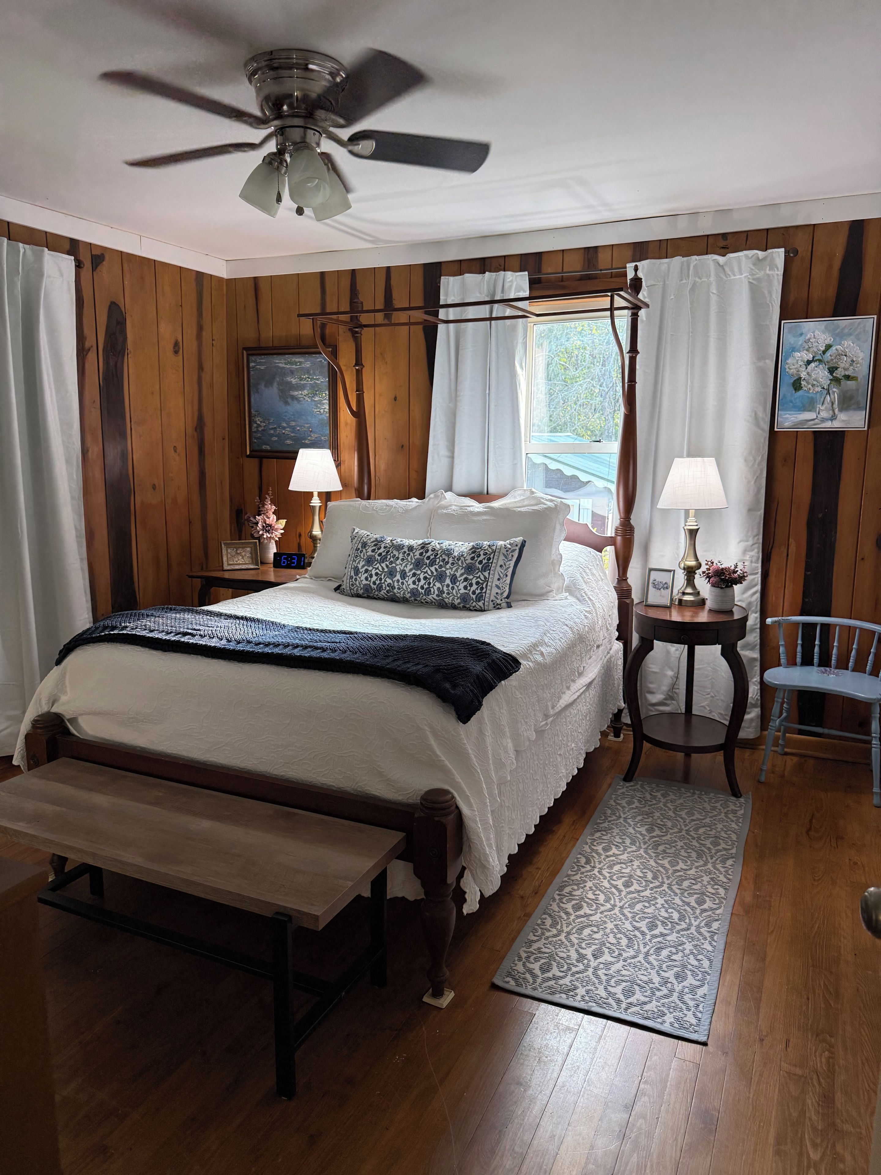 A bedroom with a wooden floor, a bed with a white bedspread, a bench, a bedside table with a lamp, a chair, and a ceiling fan.
