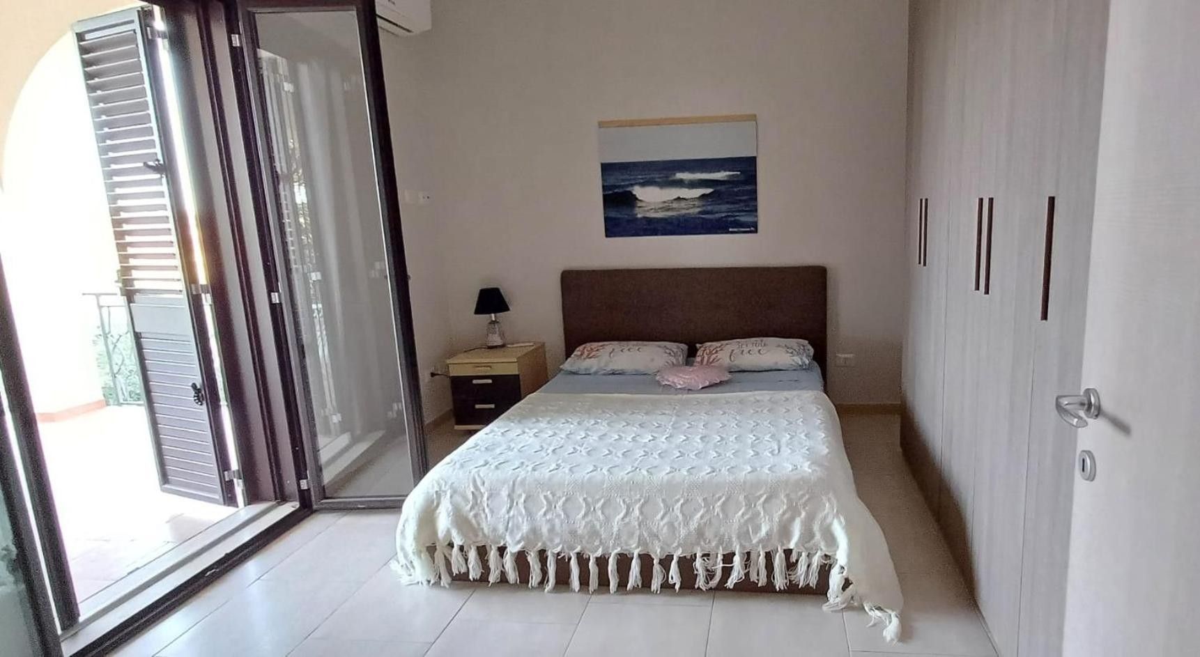 A bedroom with a bed, a nightstand, a lamp, a painting, and a sliding glass door leading to an outdoor area.