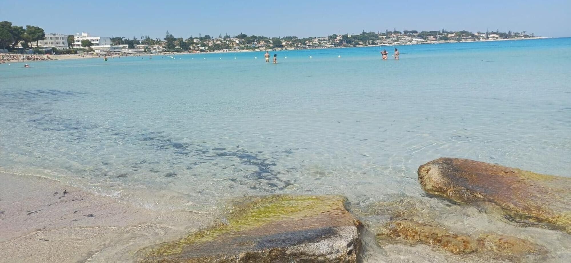 Une plage à l’eau claire, avec des rochers au premier plan et une ville en arrière-plan.