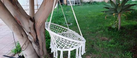 A white macrame hanging chair suspended from tree branches in a garden area with grass and a small plant.