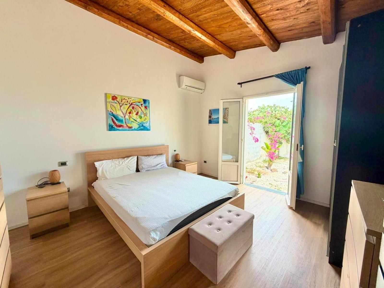 Une chambre avec un plafond en bois, un lit avec un matelas blanc, une table de chevet, un petit tabouret et une porte-fenêtre coulissante donnant sur un espace extérieur verdoyant.