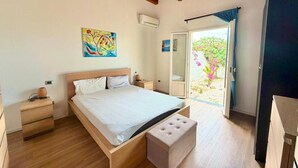 Quarto com teto de madeira, cama com colchão branco, mesa de cabeceira, um banquinho pequeno e porta de vidro deslizante que dá para uma área externa com vegetação.