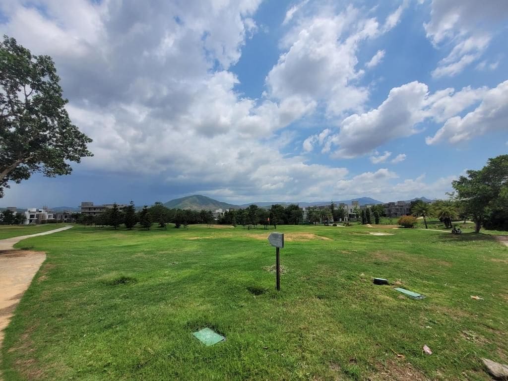An open grassy area with a signpost, a dirt path, and buildings in the distance under a partly cloudy sky.