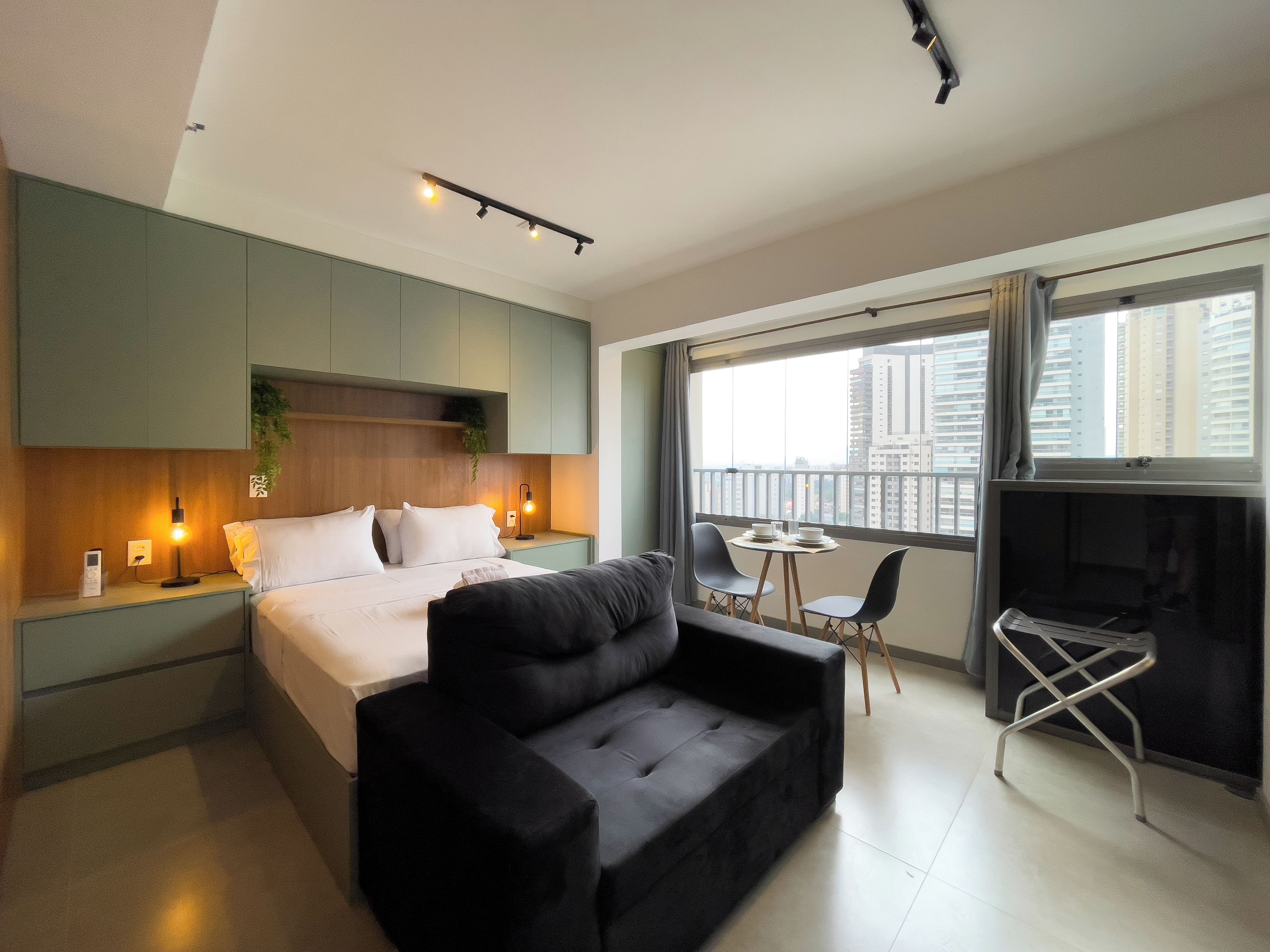 A modern hotel room with a bed, a black sofa, a small dining table, and a balcony with a city view.