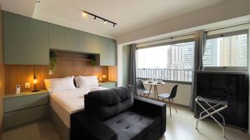 Une chambre d’hôtel moderne avec un lit, un canapé noir, une petite table à manger et un balcon offrant une vue sur la ville.