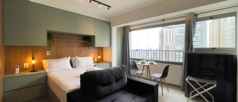 A modern hotel room with a bed, a black sofa, a small dining table, and a balcony with a city view.