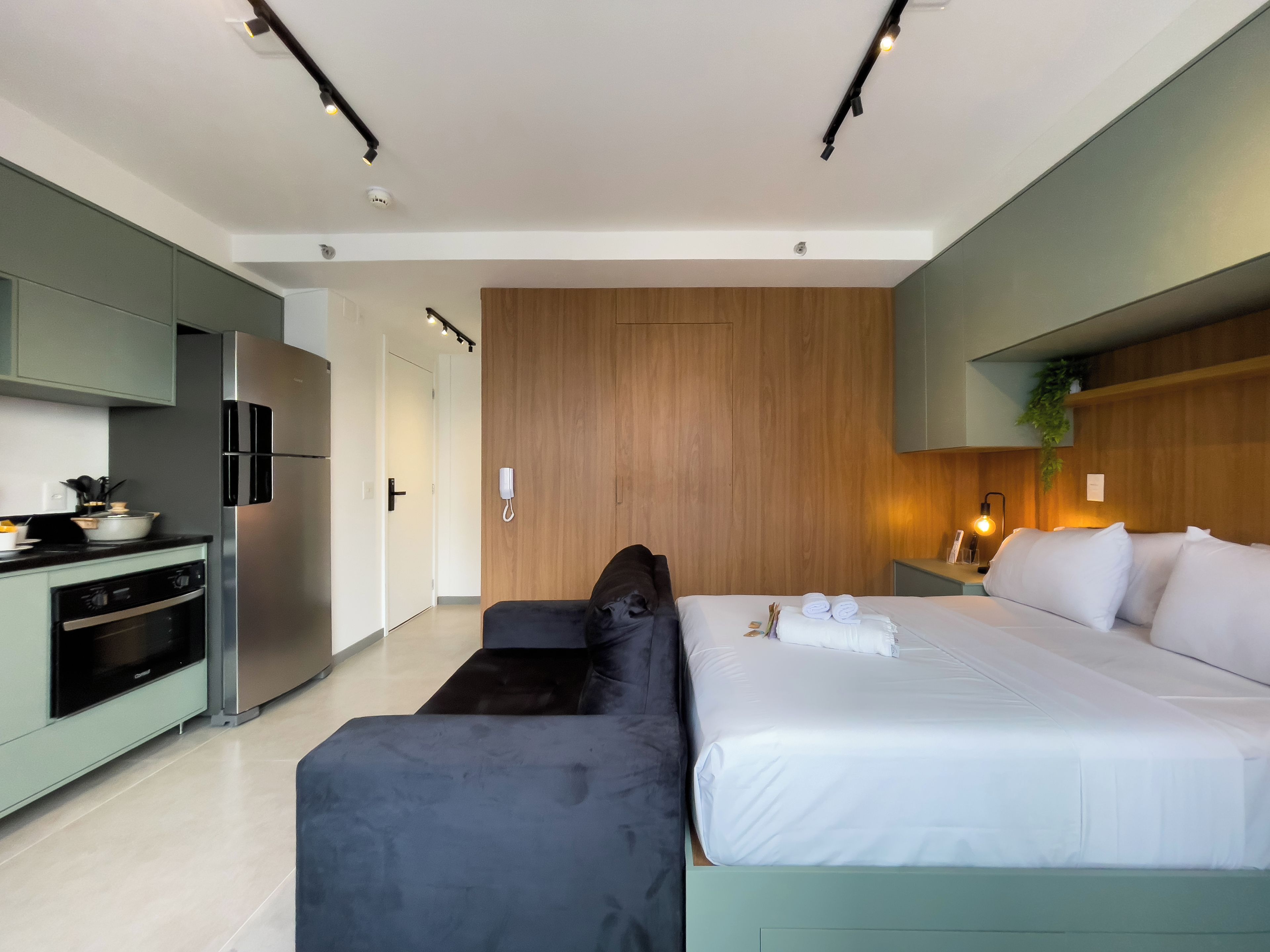 A modern hotel room with a kitchenette, a bed with white linens, a dark-colored sofa, and a wooden accent wall.