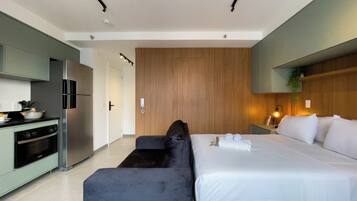 Une chambre d’hôtel moderne dotée d’une kitchenette, d’un lit avec du linge de maison blanc, d’un canapé de couleur foncée et d’un mur décoratif en bois.