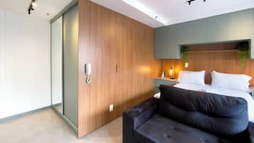 A modern bedroom with a wooden wardrobe, a black sofa, a bed with white linens, and a bedside table with a lamp.