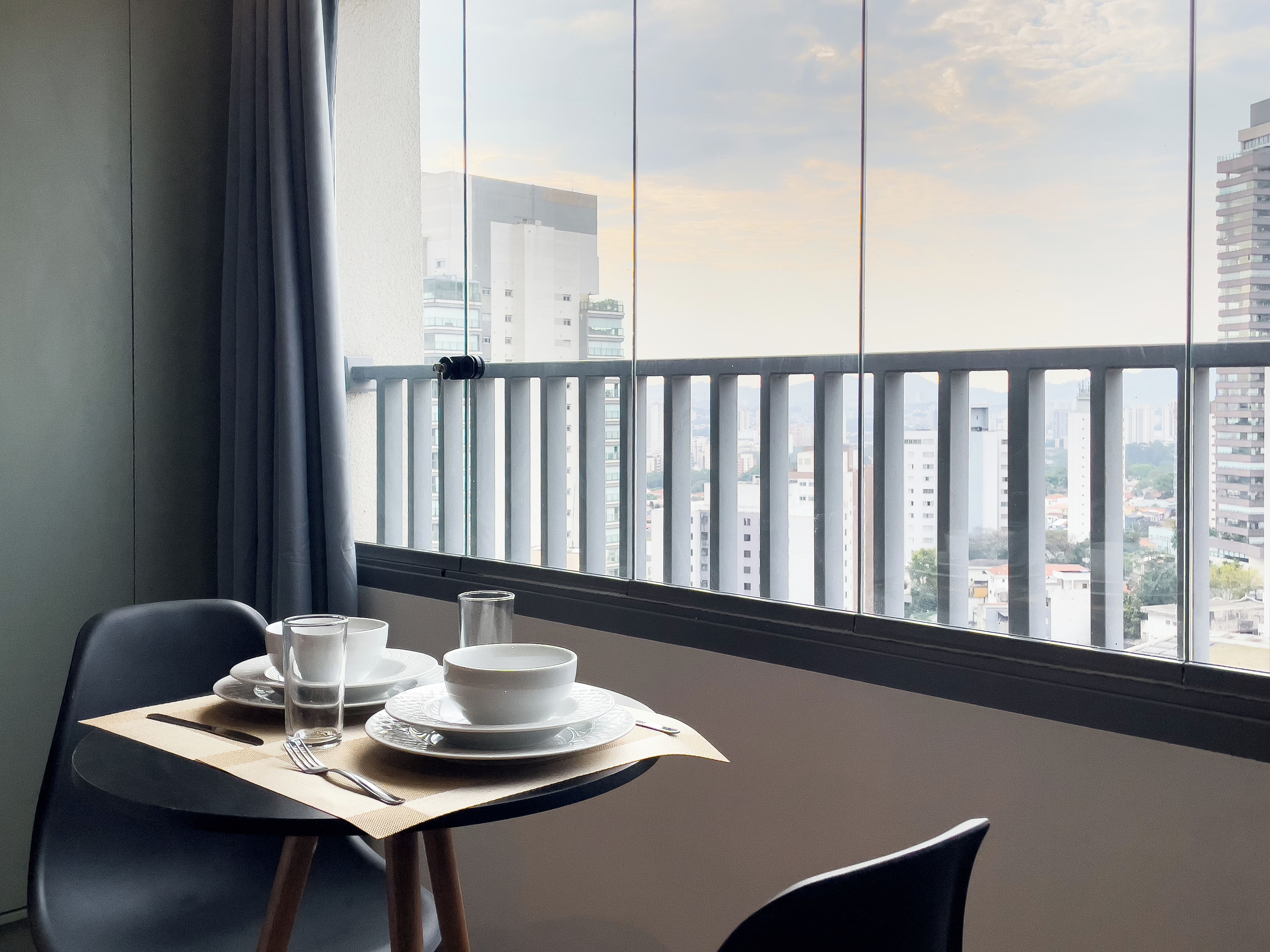 A dining area with a table set for two, a window with a view of city buildings, and a balcony railing.