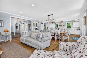 A spacious living room with a sofa, a dining area with a table and chairs, and a kitchen with white cabinets and a pendant light. - Driftwood Daydream  Dog-friendly, lakefront house with beach access, dock, AC (St.Germain)