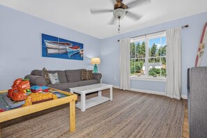 A living room with a sofa, a coffee table, a painting of boats, a window with curtains, and a ceiling fan. - Driftwood Daydream  Dog-friendly, lakefront house with beach access, dock, AC (St.Germain)