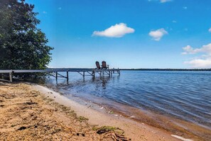 A beach with a wooden pier extending into calm water, two chairs on the pier, and a tree on the left.