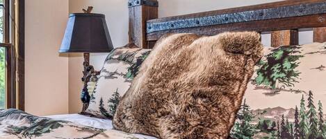 Un lit avec une couvre-lit à motifs de forêt, une tête de lit en bois et une lampe avec un abat-jour noir.