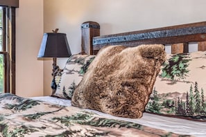 Uma cama com colcha decorada com tema de floresta, cabeceira de madeira e abajur com cúpula preta.