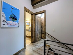 A hallway with a wooden door leading to a bathroom, a staircase with a metal handrail, and a framed picture of a ski lift on the wall. - NEW | Updated | Forest Views | Free Shuttle (Keystone)
