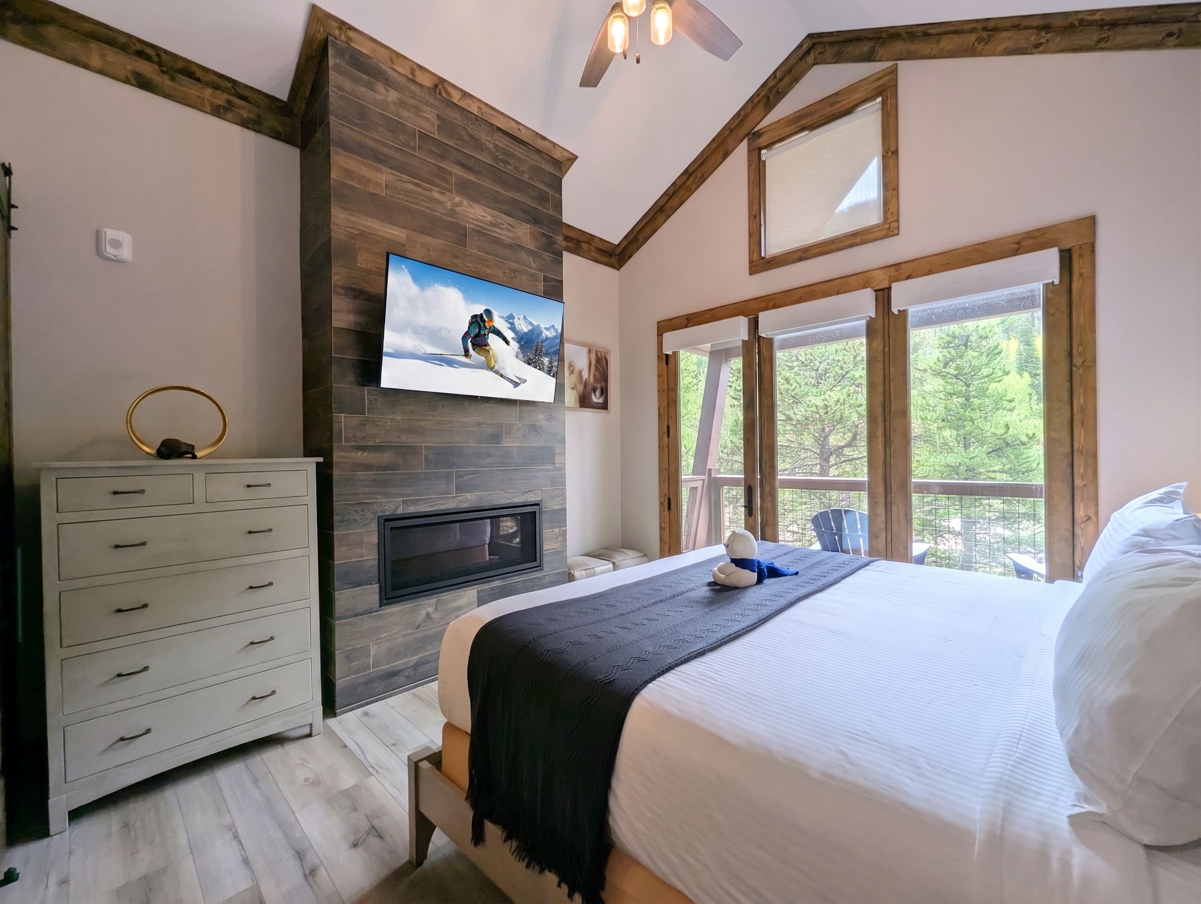 Un dormitorio con cama, cómoda, chimenea, televisor y ventanas grandes.