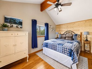 A bedroom with a wooden ceiling, a bed with a blue and white checkered bedspread, a white dresser, a window with blue curtains, and a ceiling fan. - Emberwood | Hot Tub, 2 Fireplaces, Sleeps 9 (Heber)