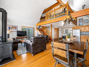 A spacious living area with a fireplace, leather sofa, dining table, and a staircase leading to an upper level. - Emberwood | Hot Tub, 2 Fireplaces, Sleeps 9 (Heber)