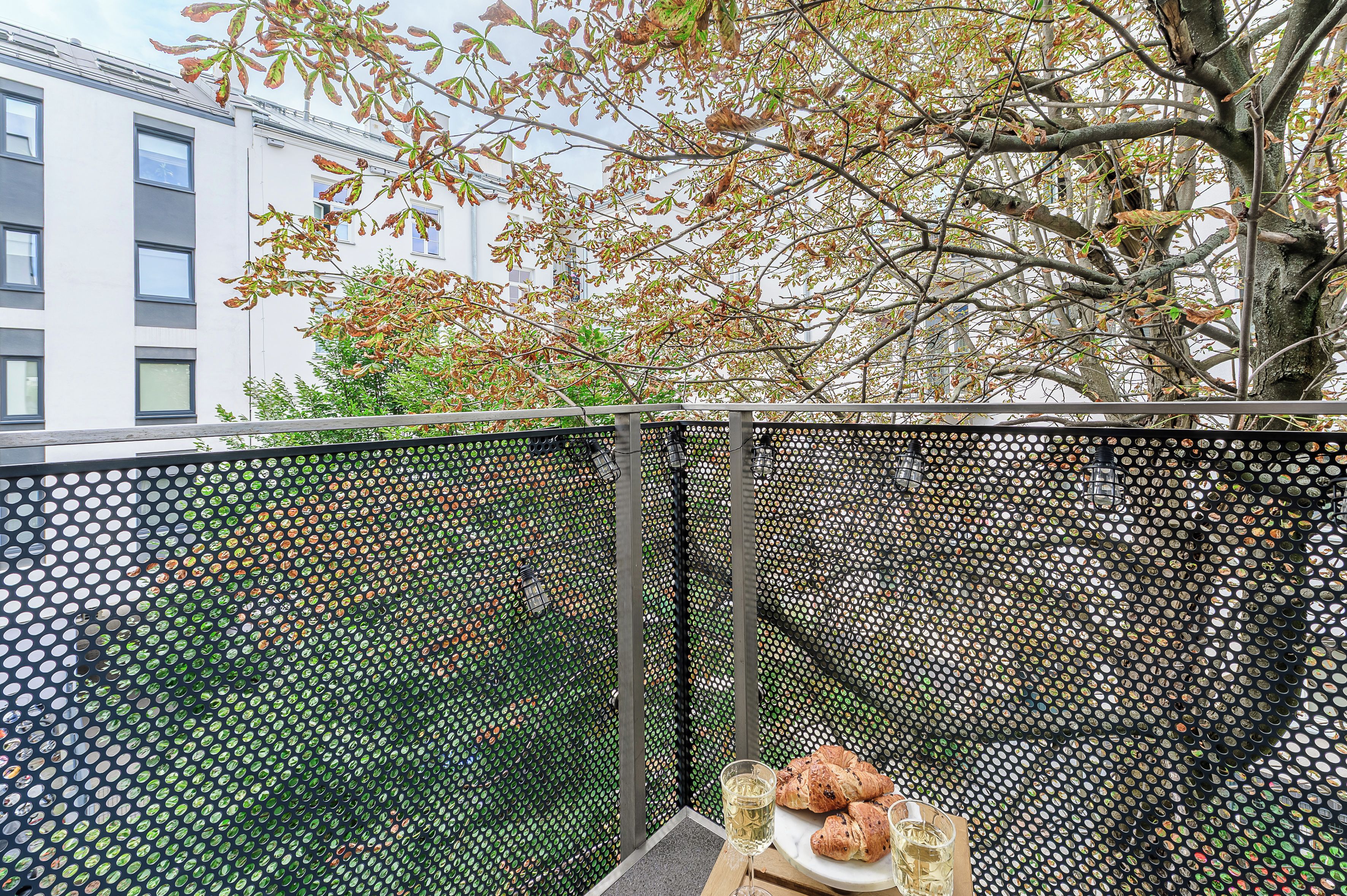 Un balcon avec une balustrade en métal, un arbre aux feuilles d’automne et une table avec des croissants et un verre de vin blanc.