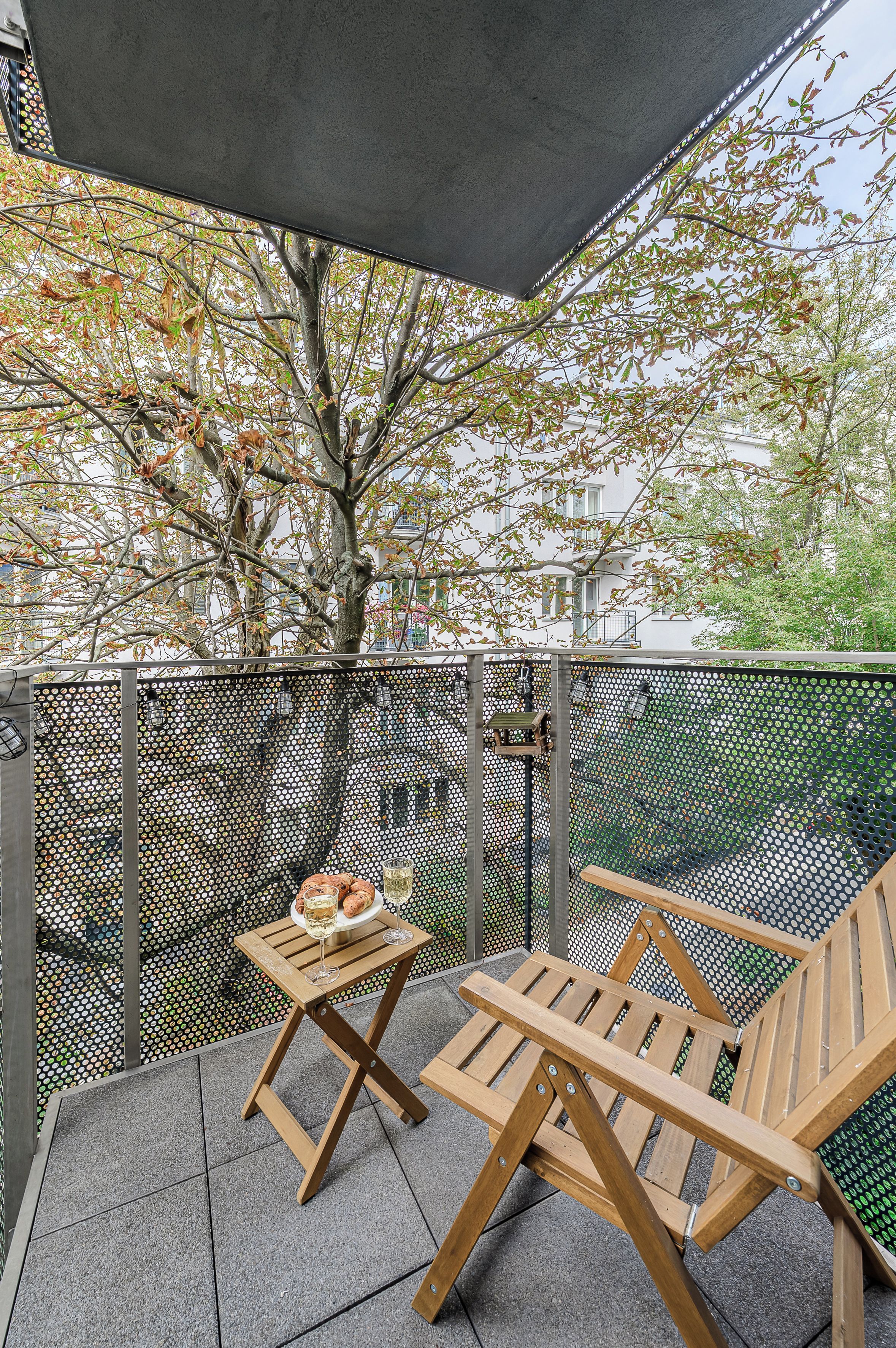 A balcony with a table and two chairs, a tree, and a building in the background.
