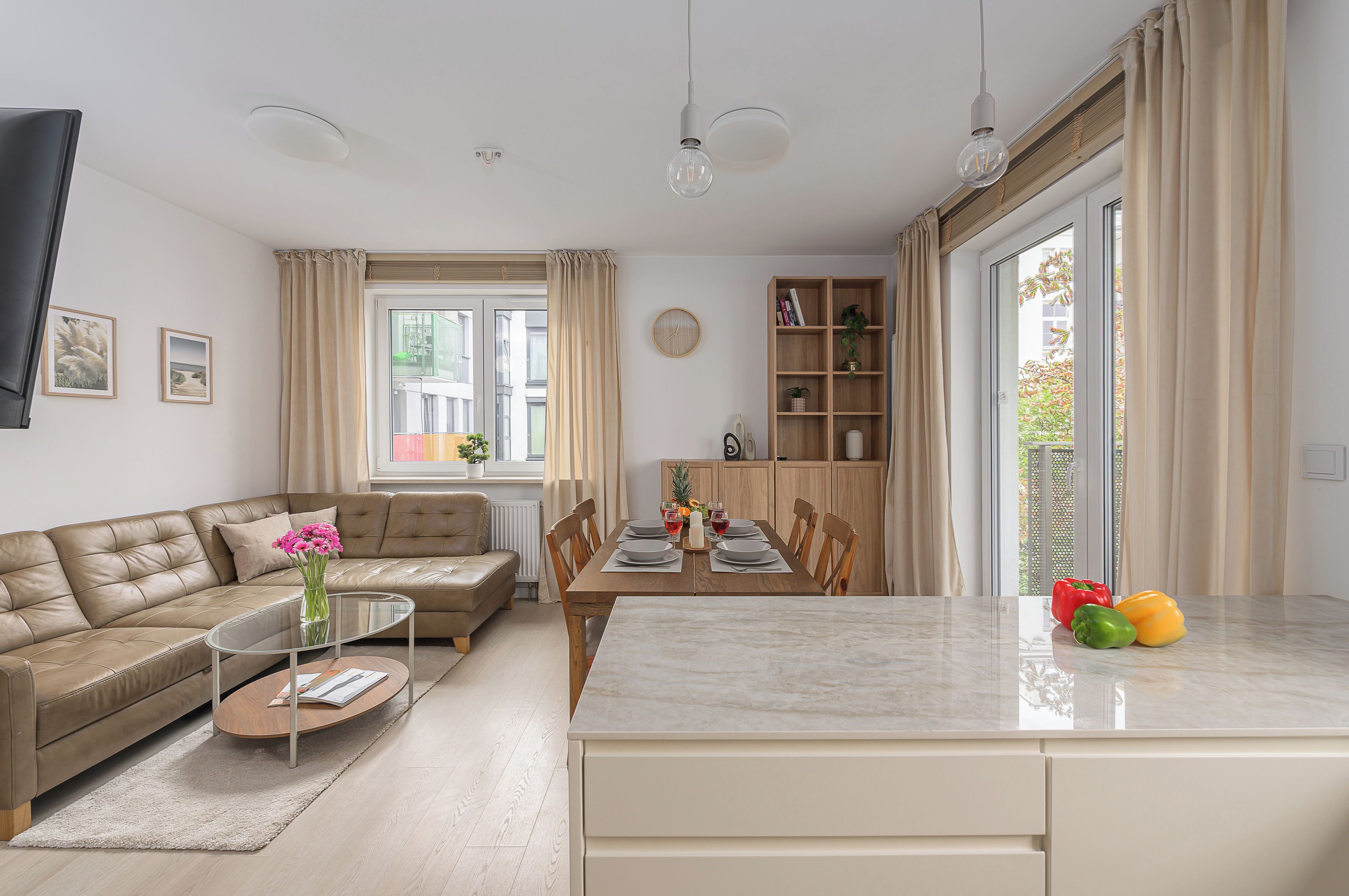A modern living room with a sofa, a dining table set for two, and a kitchen area with a wooden cabinet and a window.