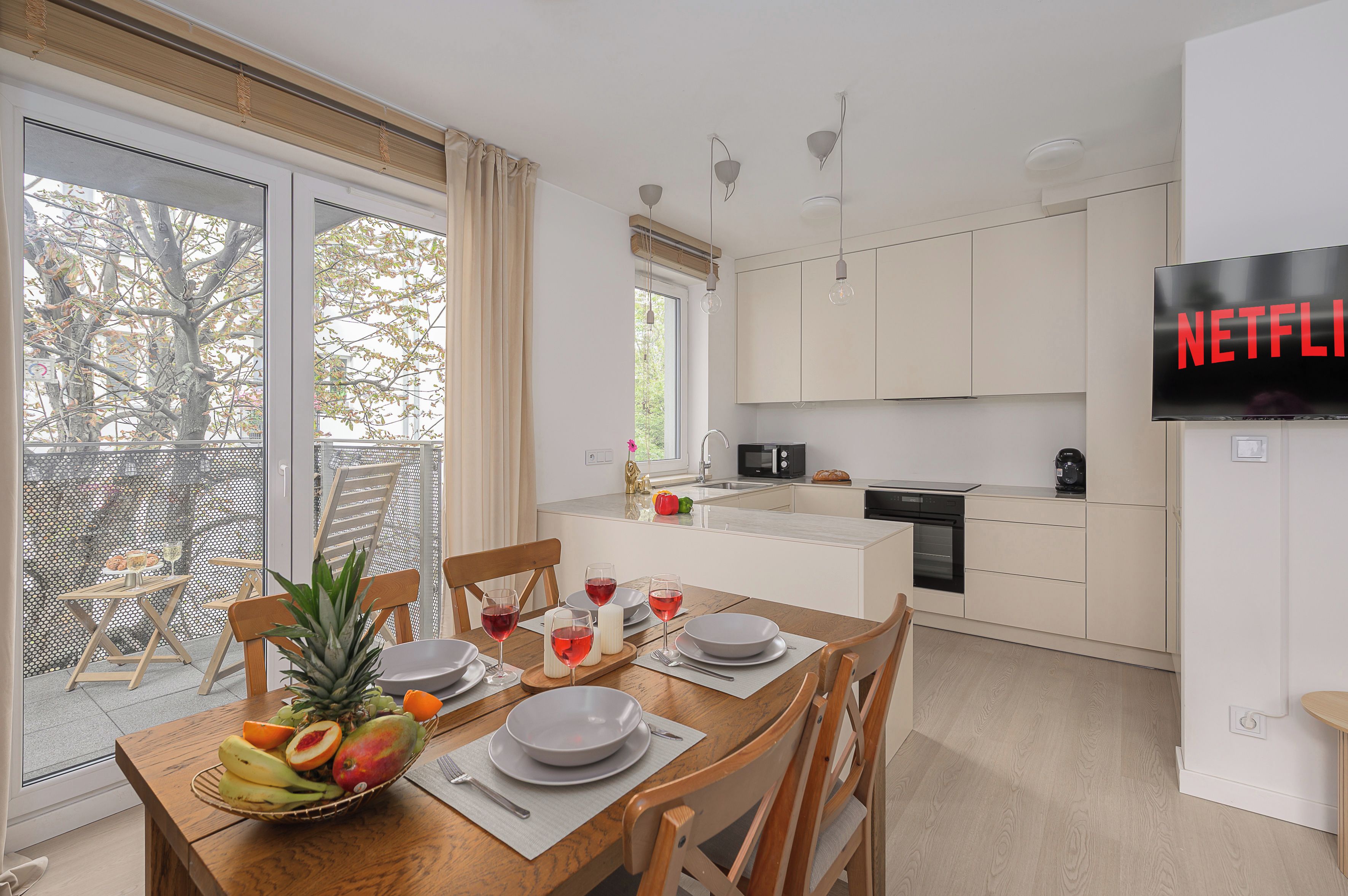 A modern kitchen with a dining area, a TV screen displaying 'NETFLIX', and a view of trees through the windows.