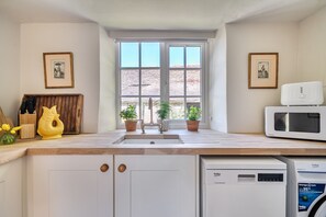 Una cocina con un lavaplatos, un microondas y un lavavajillas. Hay dos macetas en el alféizar de la ventana y un armario de madera con un bloque de cuchillos.