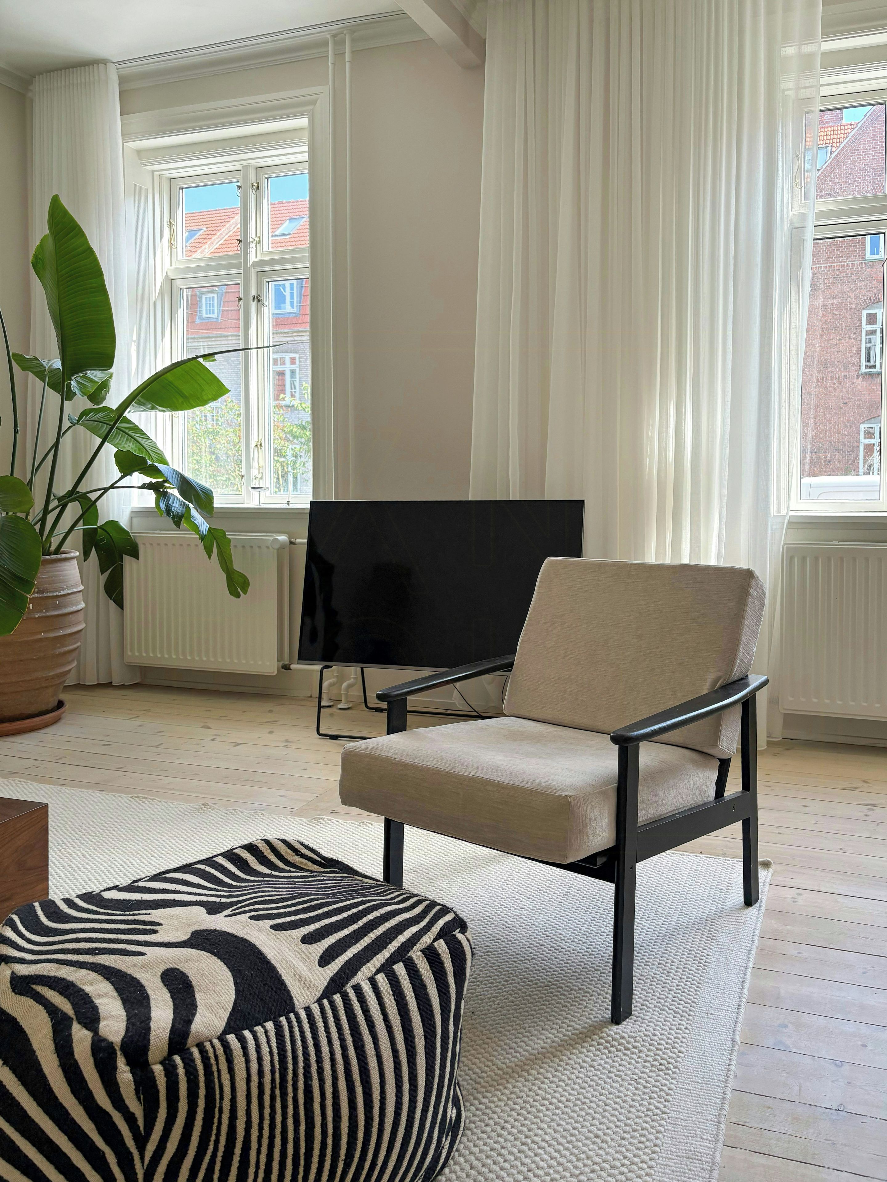 A living room with a large window, a television, a beige armchair, a zebra-patterned ottoman, and a potted plant.