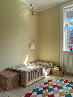 Quarto de criança com berço, tapete colorido e janela vista para edifícios.