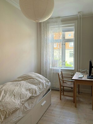 Ein Schlafzimmer mit einem Bett, einem Schreibtisch mit Stuhl, einem Fenster mit Vorhängen und einer Pendelleuchte.
