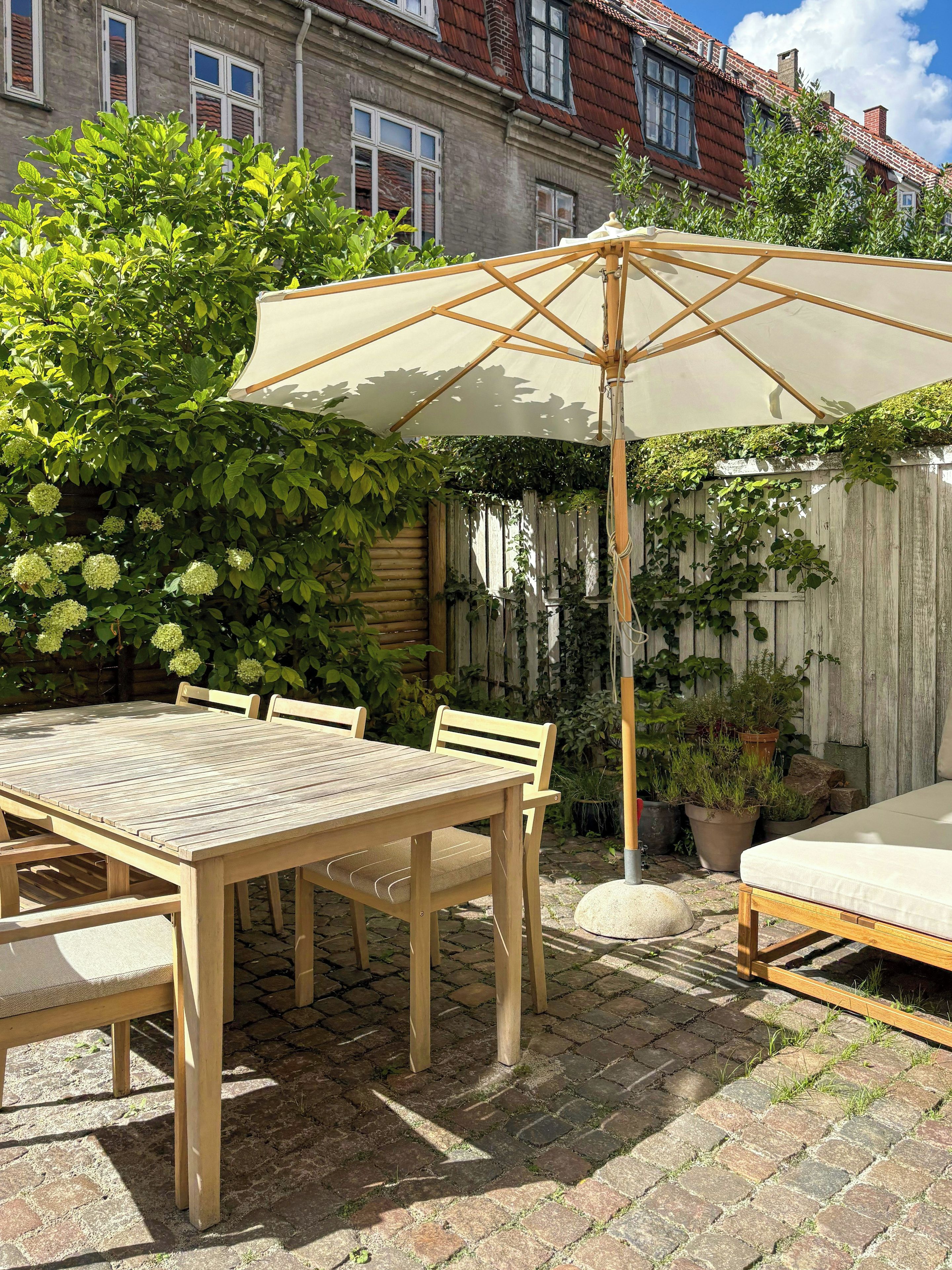 A wooden patio table with chairs, a white umbrella, and a mattress on the ground.