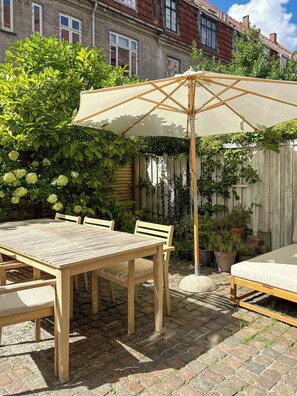 Ein Holztisch mit Stühlen, ein weißer Sonnenschirm und eine Matratze auf dem Boden.
