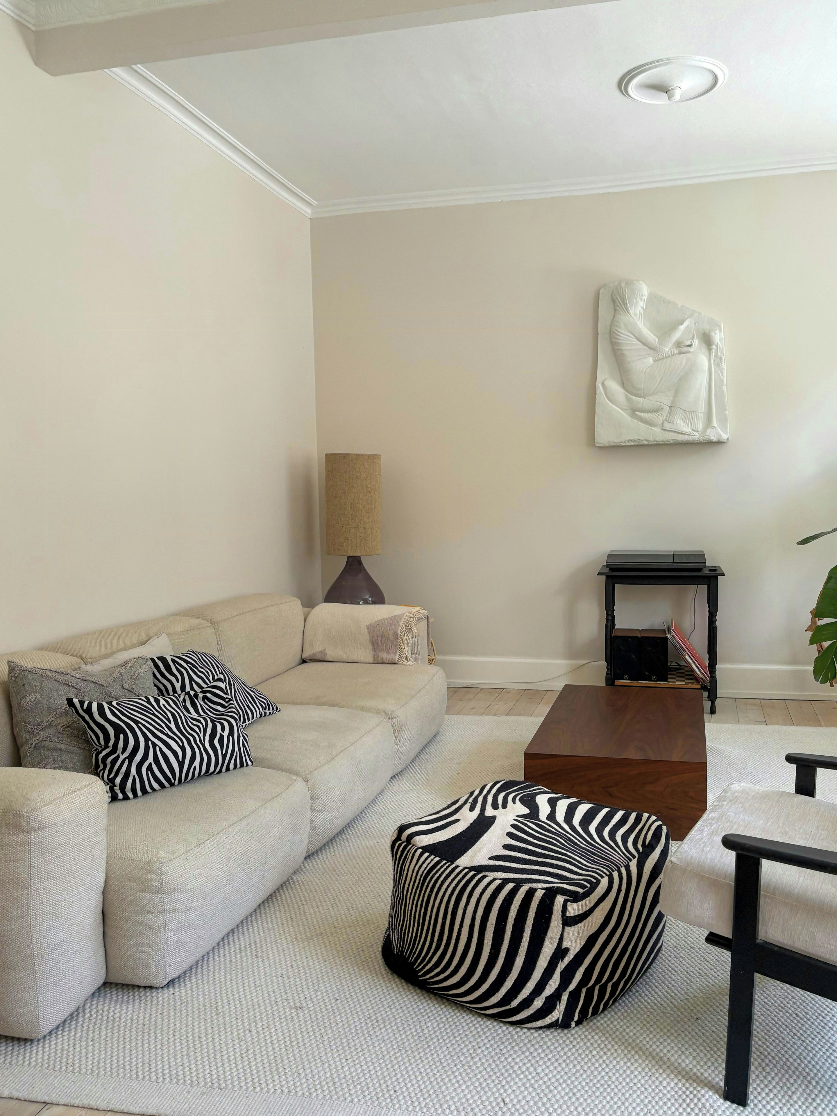 En stue med en beige sofa, puder med zebra-mønster, et træsofabord og en vægmonteret skulptur.